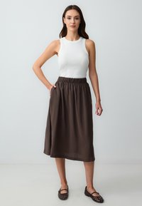 Gonna marrone a lunghezza ginocchio con vita elasticizzata, abbinata a una maglietta bianca senza maniche e ballerine intrecciate marrone, su uno sfondo neutro.