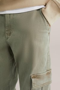 Osoba v světle zelených cargo kalhotách a béžovém žebrovaném svetru, s rukou v kapse, detailní záběr na pas a stehna.