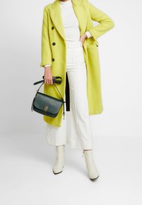 Lungo cappotto verde lime con bottoni neri, abbinato a pantaloni bianchi a vita alta e stivali alla caviglia color crema. Una borsa verde scuro completa l'outfit.