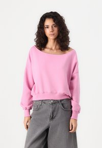 Felpa oversized rosa in un tessuto morbido, caratterizzata da un ampio scollo e polsini elastici, abbinata a pantaloni di jeans grigi a vita alta.