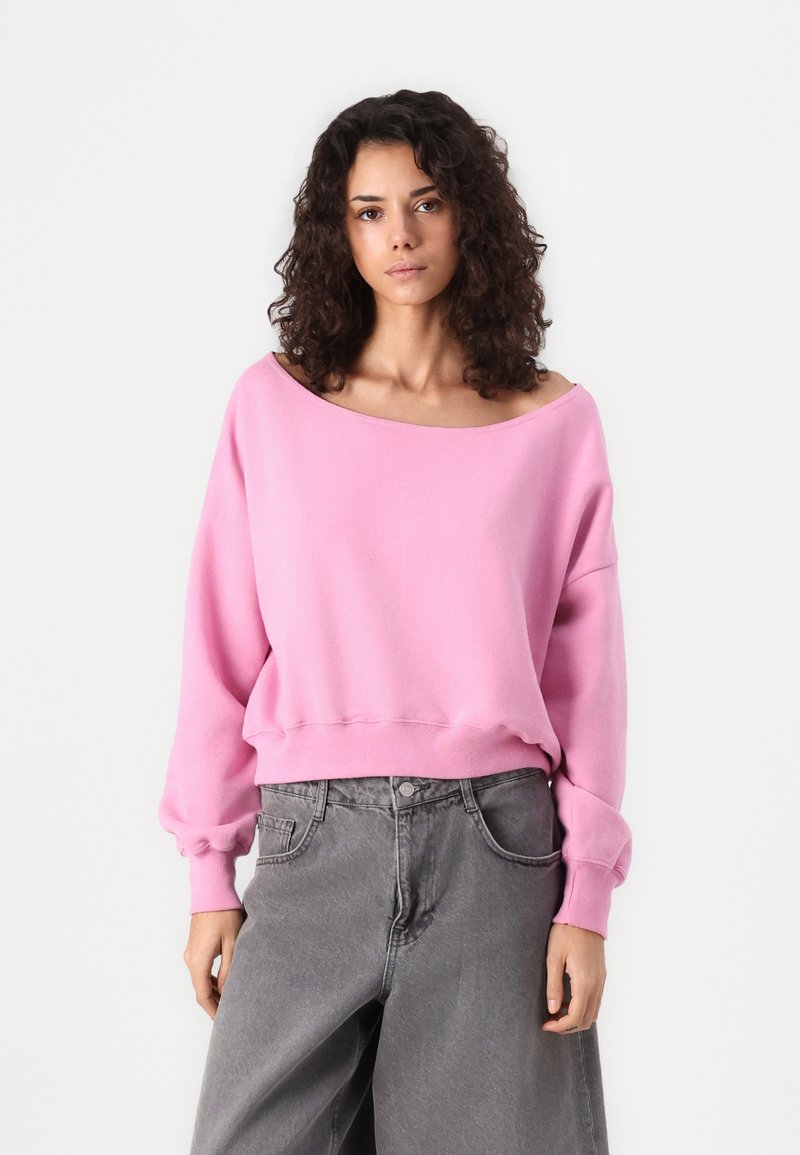 Felpa oversized rosa in un tessuto morbido, caratterizzata da un ampio scollo e polsini elastici, abbinata a pantaloni di jeans grigi a vita alta.