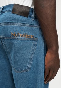 Πέτο τζιν με ταμπά κέντημα "No Problemo", μαύρο δερμάτινο ετικέτα στη ζώνη, φορεμένο από άτομο με σκουρόχρωμο δέρμα και άσπρο πουκάμισο.