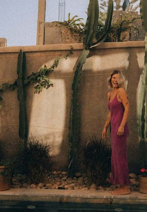 Różowa sukienka bez rękawów z cienkimi ramiączkami, z lejącym się materiałem i luźnym krojem. W tle znajdują się wysokie kaktusy i dekoracyjne rośliny na tle ściany.