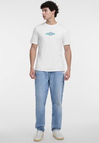 Baltas medvilninis marškinėliai su ryškiu "Guess Jeans" logotipu, derinami su šviesiai mėlynais nusidėvėjusiais džinsais ir baltais sportbačiais.