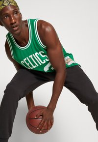 Grünes Basketball-Trikot mit weißen Akzenten und "CELTICS" in großen Buchstaben bedruckt, kombiniert mit schwarzen Sporthosen. Spieler hält einen Basketball.