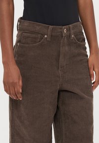Bruine corduroy broek met een ribbelstructuur, voorzien van voorzakken, een knoopsluiting en een aansluitende taille, weergegeven van de taille naar beneden.