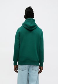 Grøn hættetrøje med løs pasform, der har ribstrikkede ærmer og kant. Båret med lyseblå denimjeans.