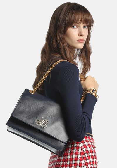 Femme aux longs cheveux bruns portant un pull marine et une jupe à carreaux rouges, tenant un sac à main en cuir marine avec une chaîne dorée sur l'épaule.