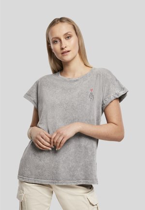 Femme blonde portant un t-shirt gris aux manches retroussées avec un petit dessin de main tenant un cœur rouge sur la poitrine, associée à un pantalon cargo clair.