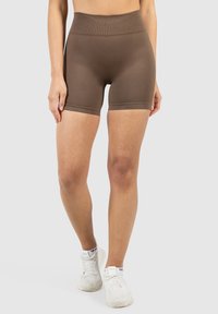 Das Modell trägt braune hochgeschnittene Shorts, kombiniert mit weißen Sneakers, und steht vor einem schlichten, hellen Hintergrund.