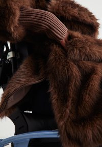 Άτομο που φοράει ένα παχύ καφέ γούνινο παλτό και πλεκτό πουλόβερ με ραβδώσεις, καθισμένο σε αναπηρικό καροτσάκι με ορατό μπλε σκελετό.