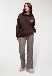 Felpa marrone con tasca frontale e coulisse, abbinata a pantaloni a gamba larga grigi a quadri e scarpe platform grigio chiaro.