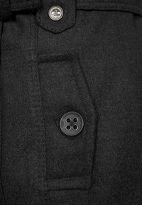 Schwarzes Wollkleidungsstück mit einer Fronttasche und einem runden schwarzen Knopf. Der Metallknopf zeigt den eingravierten Markennamen. Weiche Textur und klare Linien.