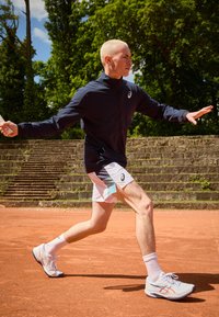 Atleta con chaqueta azul marino y pantalones cortos multicolores en una cancha de tenis de arcilla, llevando zapatillas claras y calcetines blancos, concentrado en una raqueta de tenis.