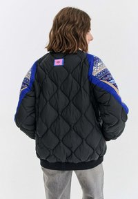 Prešívaná čierna bunda s modrými rukávmi, ktorá má geometrický vzor a ružovú nášivku na chrbte. Hladký lem a manžety.