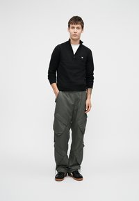 Sweater preto com fecho, calças cargo verdes com bolsos laterais e sapatilhas pretas com solas castanhas. Modelo está encostado a um fundo branco liso.