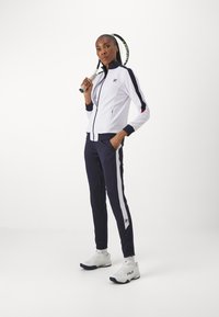 Valkoinen vetoketjullinen urheiluva jacket, jossa on laivastonsinisiä yksityiskohtia, yhdistettynä laivastonsinisiin verkkareihin, joissa on valkoiset raidat. Valkoiset urheilukengät viimeistelevät asustuksen.