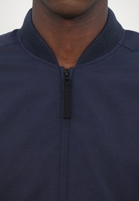 Námornícky modrá bomber jacket s rebrovaným golierom, predným zipsom a hladkou textúrou látky. Minimalistický dizajn bez viditeľných vzorov alebo akcentov.