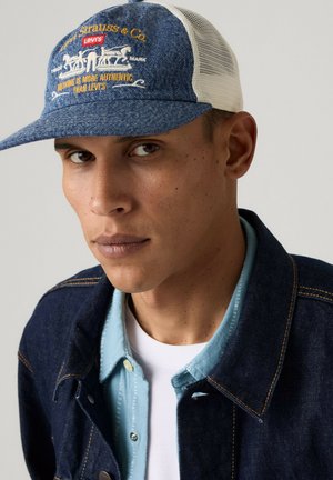 Mann trägt eine blaue Levi's Jeanskappe mit weißem Netzrücken, geschichtete Jeanshemden, schaut leicht seitlich mit neutralem Gesichtsausdruck.