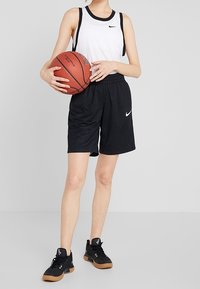 Pantalones cortos deportivos negros con cintura elástica, combinados con una camiseta sin mangas blanca con acentos negros. Sosteniendo un balón de baloncesto, usando zapatillas negras.