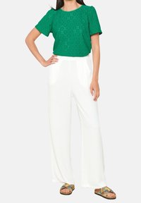 Grünes Eyelet-Oberteil mit kurzen Ärmeln, kombiniert mit weißen weiten Hosen. Braune Sandalen mit bunten Akzenten.