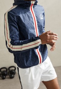 Námořnická větrovka s bílými a červenými pruhy, se znakem a celoplošným vzorem, v kombinaci s bílými šortkami pro sportovní vzhled.