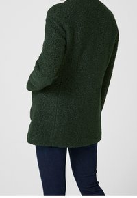 Persona vestida con un abrigo de lana texturizada verde oscuro y de mangas largas, combinado con jeans ajustados azul oscuro, de pie contra un fondo liso.