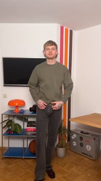 Ung mand med tatoveringer står i et rum med et vægmonteret TV, orange lampe, basketball, plante og en kuffert mærket "Tropicana".