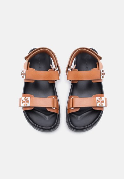 Καφέ δερμάτινα σανδάλια με δύο ρυθμιζόμενους ιμάντες και ασημένιες αγκράφες σε μαύρες σόλες, απεικονίζονται από πάνω σε λευκό φόντο.