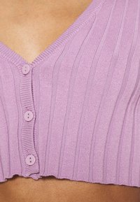 Närbild av en ribbad, lavendelfärgad kortärmad cardigan med tre knappar knäppta över bar hud.