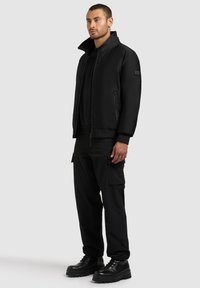 Jachetă bomber neagră cu guler înalt, fermoar frontal și buzunare laterale, purtată împreună cu pantaloni cargo negri și cizme masive negre.