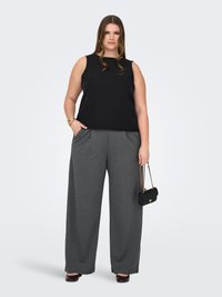 Vrouw met zwarte mouwloze top, grijze broek met wijd uitlopende pijpen, bordeauxrode schoenen, houdt een zwarte gewatteerde tas met gouden ketting vast tegen een witte achtergrond.