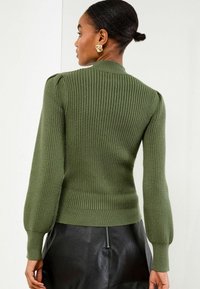 Rippstrick grüner Pullover mit hohem Kragen, Puffärmeln und eng anliegenden Bündchen, kombiniert mit einem schwarzen Lederrock.