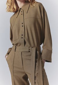 Braunes, strukturiertes Hemd mit Knopfleiste und spitzem Kragen, mit einer Brusttasche, kombiniert mit passenden hoch taillierten Hosen, die einen gebundenen Gürtel haben.