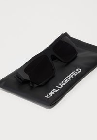 Černé sluneční brýle s matným povrchem, geometrickým rámem a tmavými skly odložené na černém pouzdře s bílým textem "KARL LAGERFELD".
