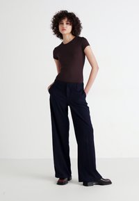 Brown fitted t-shirt paired with wide-leg navy trousers. Model stands confidently, wearing black and red block-heeled shoes. Simple background.
