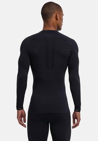 Czarna sportowa koszulka z długim rękawem o prążkowanej fakturze, dopasowanym kroju i odblaskowym detalem na mankietach. Z tyłu znajduje się panelowy wzór.