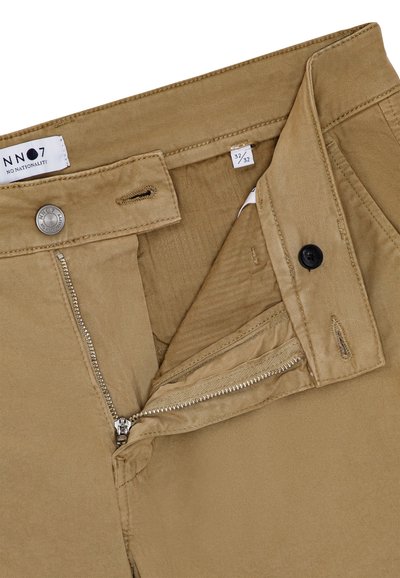 Pantalon beige avec fermeture éclair partiellement ouverte et bouton, l'étiquette indique "NN07 No Nationality", taille 32/32 pour la taille et la longueur.