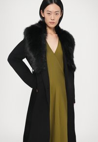 Melnā mētelis ar kažokādas apkakli, pārklāts virs zaļas kleitas. Raksturīgas īpašības ir V-veida kakla izgriezums un plūstošs dizains, kas piedāvā modernu siluetu.