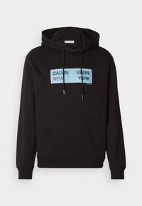 Zwarte hoodie met een kangaroo zak aan de voorkant, met een blauwe rechthoekige grafische afbeelding waarop "CALVIN KLEIN NEW YORK" in zwarte vetgedrukte letters is afgedrukt.