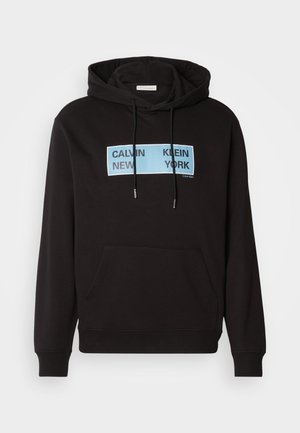 Juodas gaubtas su priekinėmis kengūros kišenėmis, su mėlynu stačiakampiu grafiku, ant kurio juodomis didžiosiomis raidėmis užrašyta "CALVIN KLEIN NEW YORK".