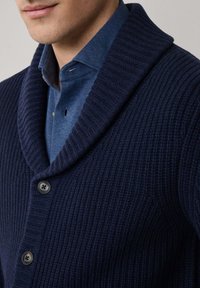 Cardigan blu navy a coste con colletto a scialle, caratterizzato da bottoni scuri e lavorazione a maglia testurizzata. Indossato sopra una camicia blu con colletto.