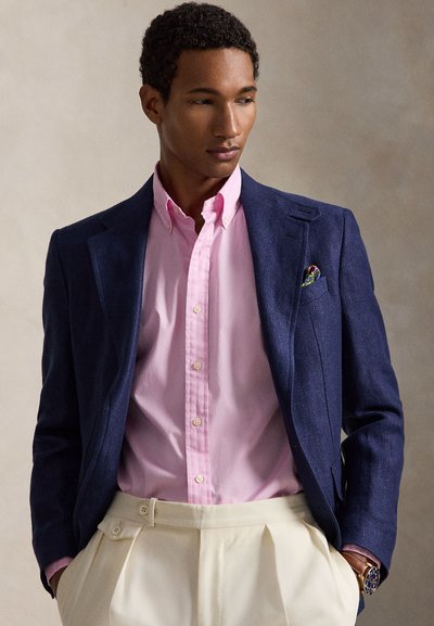 Άνδρας με ναυτικό μπλέ σακάκι, ροζ πουκάμισο, μπεζ πλισέ παντελόνι και πολύχρωμο μαντήλι στην τσέπη, με τα χέρια στις τσέπες, κοιτάζει πλάγια.