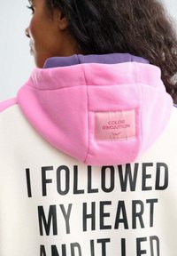 Kaptur w kremowym kolorze z wyrazistym czarnym napisem "PODĄŻYŁEM ZA SWOIM SERCEM I ON MNIE POPROWADZIŁ." Ma różowo-fioletowy kaptur i naszywkę z logo.