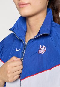 Nike vindjacka i blått, vitt och rött. Har dragkedjestängning, hög krage och broderad lejonlogotyp på vänster bröst. Texturerat material.