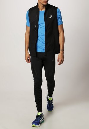 Veste noire sans manches portée sur une chemise à manches courtes bleue, assortie à un pantalon noir et des chaussures de sport bleues avec des accents verts.