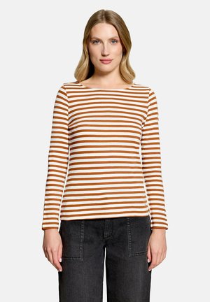 Femme aux cheveux blonds portant une chemise blanche à manches longues avec des rayures horizontales marron et un pantalon noir, debout devant un fond uni.