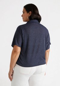 Top blu navy a maniche corte con un leggero luccichio, caratterizzato da un collo a lupetto e una vestibilità rilassata, abbinato a pantaloni bianchi. La texture appare liscia.