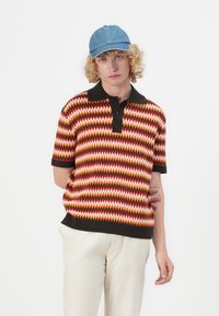 Kortermet poloshirt i oransje, rød og hvit zigzag-mønster, med svart krage og tre-knappers knepping, parret med kremfargede bukser og en denim-cap.