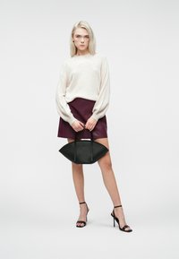 Młoda kobieta w kremowym swetrze, bordowej skórzanej spódnicy, czarnych sandałach na obcasie, trzymająca czarną półokrągłą torebkę na tle białym.